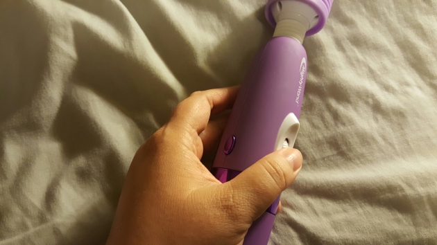 A closeup of a woman's hand grasping the body of the purple mighty wand vibrator, with her thumb on the wheel control, shown angled to the right side.