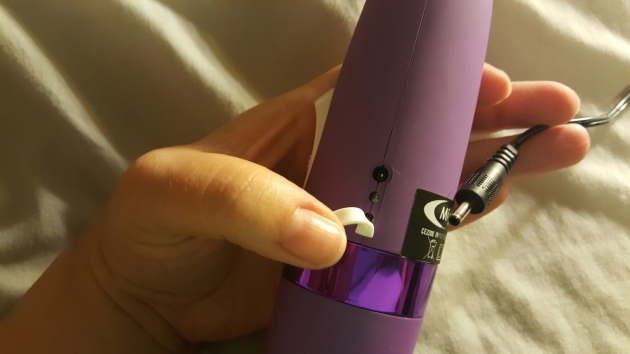 A woman's hand holding the body of the purple mighty wand vibrator. Her thumb is pulling back a small white rubber flap to reveal the charging port and two unlit LED indicators below it. She holds the wire and charging plug beside the body, grasped between her middle and index fingers to show the plug beside the port.
