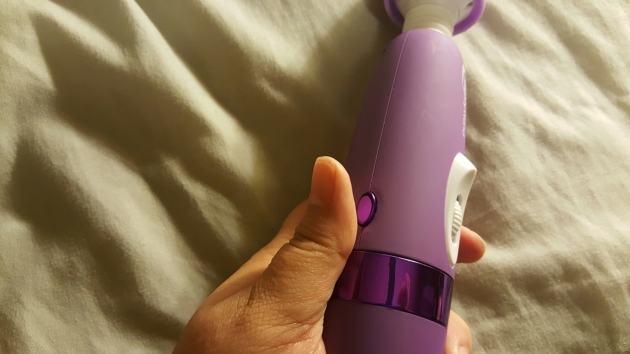 A woman's hand holding the body of the purple mighty wand vibrator. Her thumb is raised just above a shiny oval purple button on the left side of the body, about to press it.